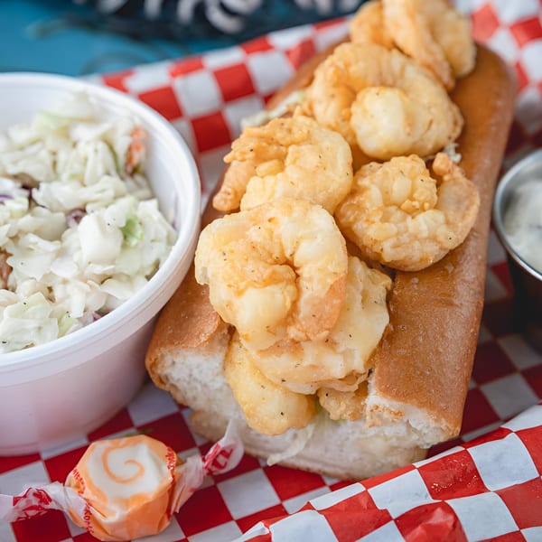 Fried shrimp po'boy with coleslaw, sauce, and a taffy candy on a red checkered tray.