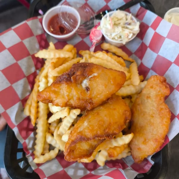 Fried fish and crinkle-cut fries in a basket with ketchup and coleslaw on a red and white checkered liner.
